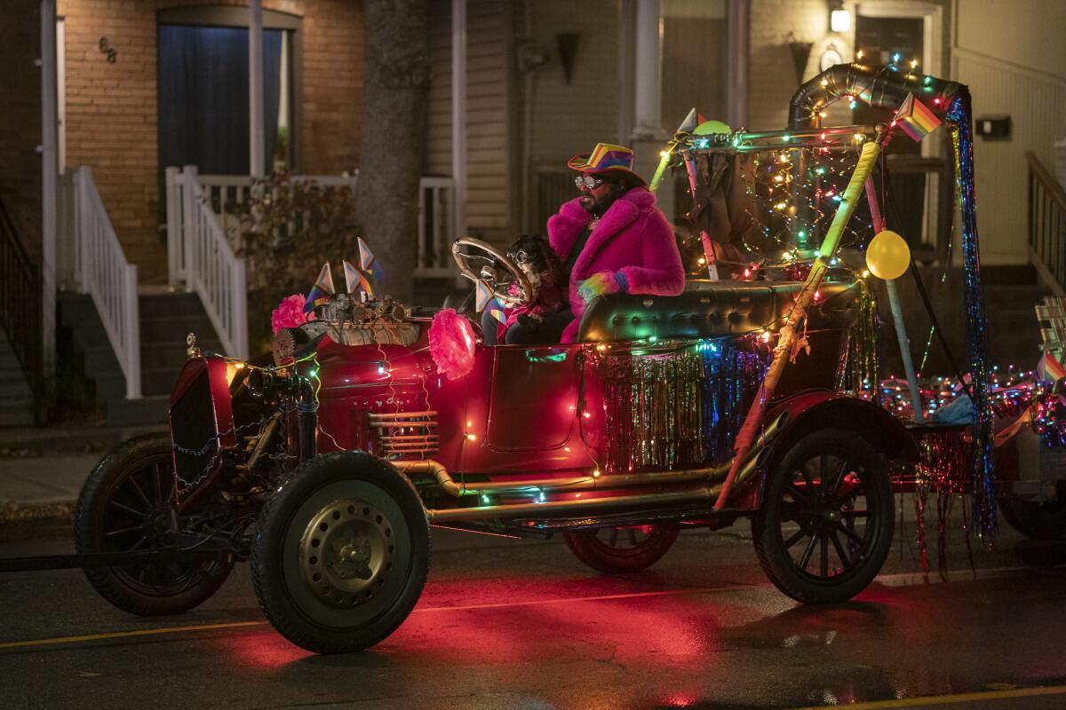 A man in a pink furry coat and hat drives an old car covered in string lights.