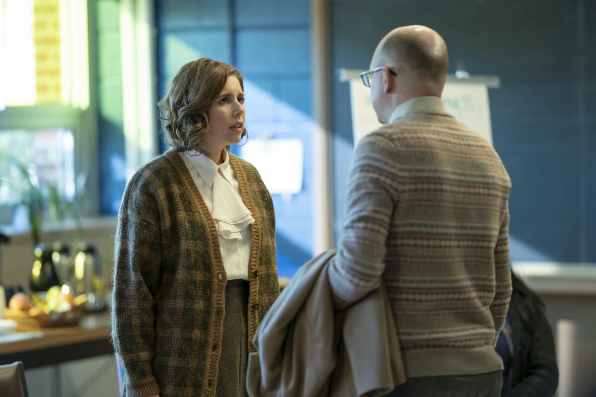 A woman in a white shirt and plaid beige cardigan looks a man in a beige sweater who is seen from behind.