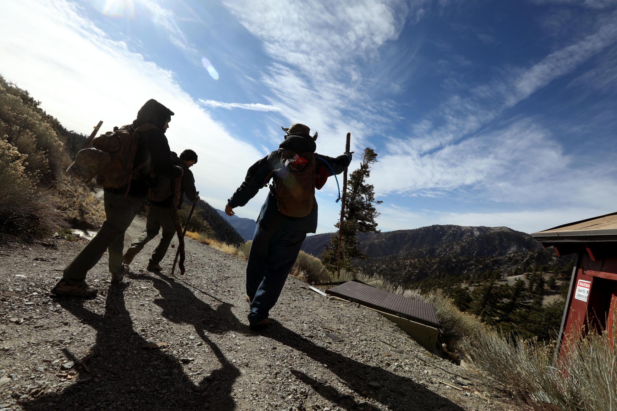 Three men walking on a dirt trail in a mountainous setting under blue skies.