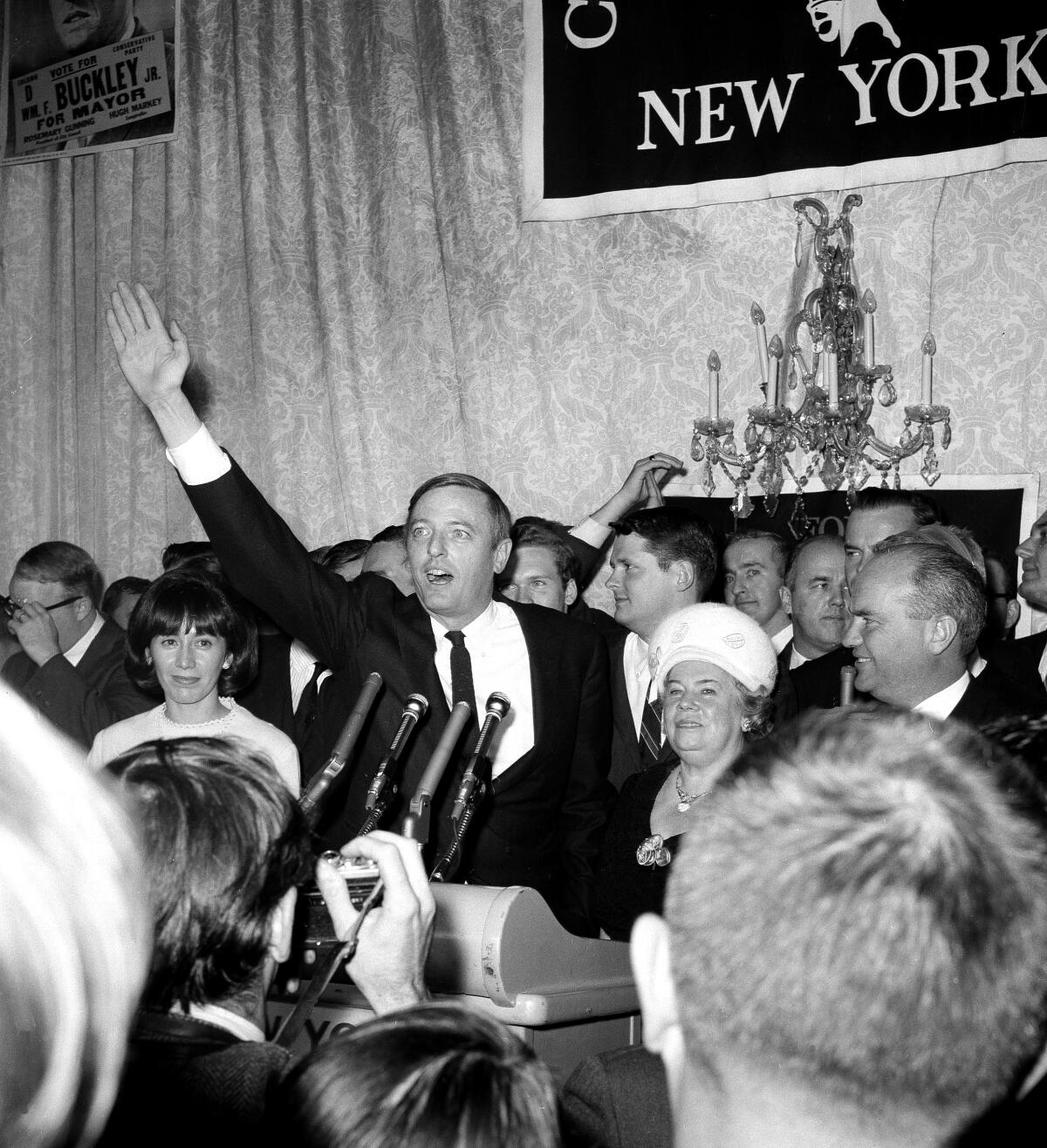 A black-and-white photo of William F. Buckley Jr. in front of several microphones, waving from the front of a crowd