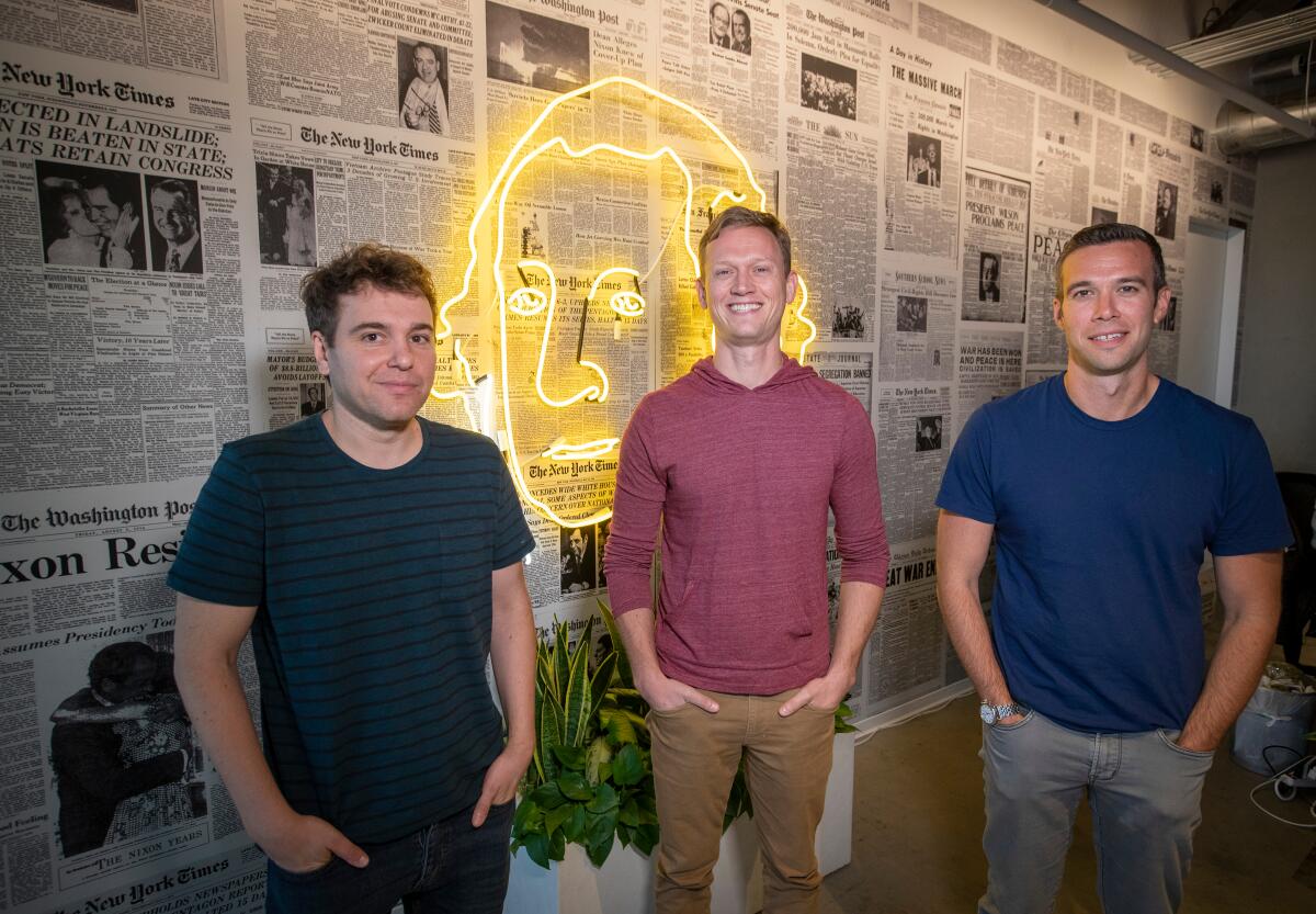 Jon Lovett, Tommy Vietor and Jon Favreau stand in front of a yellow neon image of George Washington on a wall of newspapers