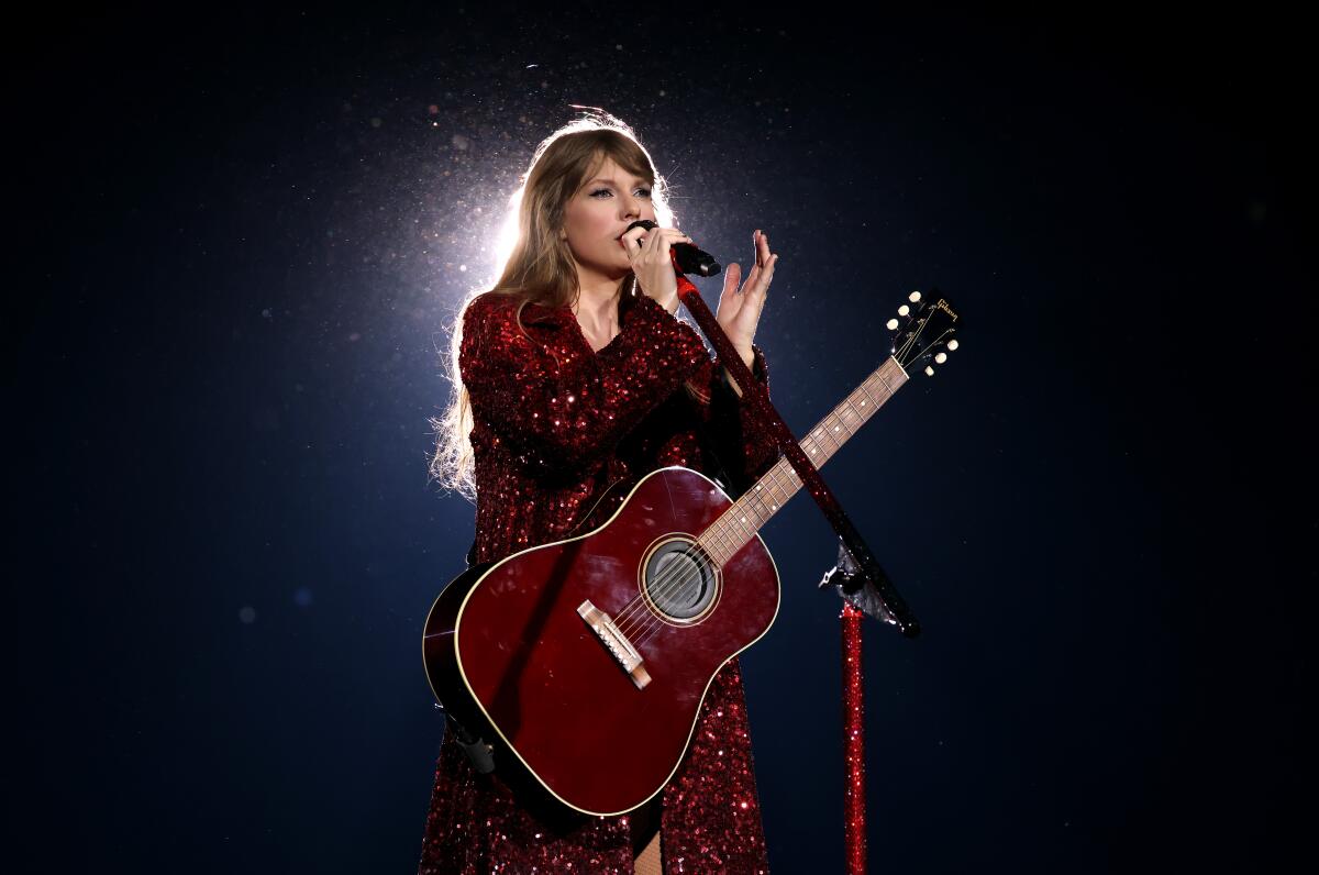 Taylor Swift standing against a dark background with an acoustic guitar, using a microphone