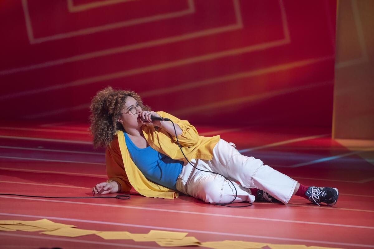 A woman in a bright yellow shirt lays down on a stage holding a microphone.