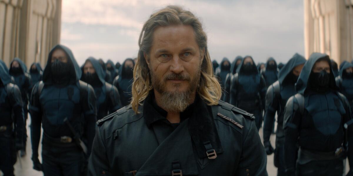 A man with long hair and a beard in a dark uniform stands in front of an army of soldiers.