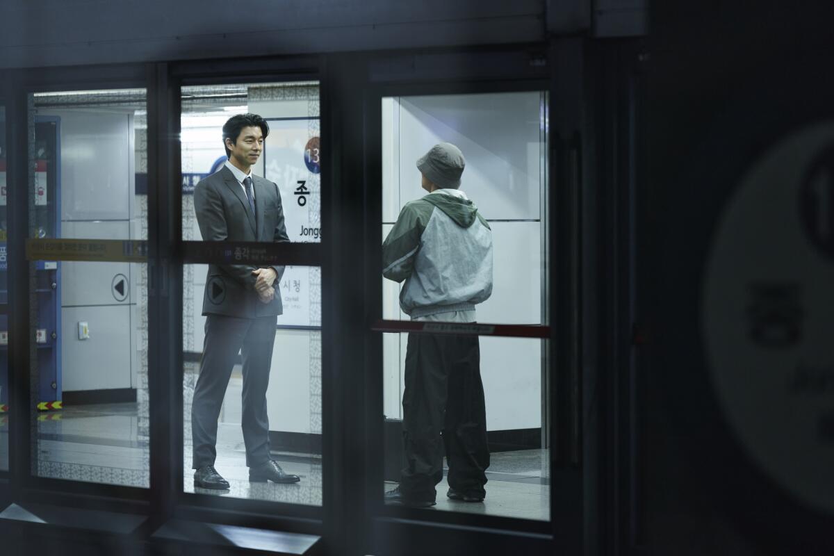 a man in a suit facing a man in a hoodie and hat in a hallway