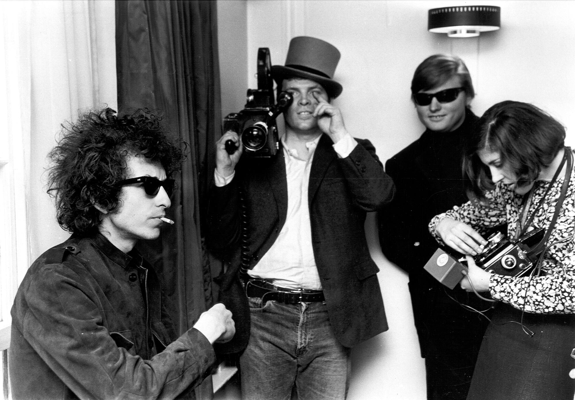 A man in sunglasses smoking near a man carrying a video camera and a man and a woman taking photographs.