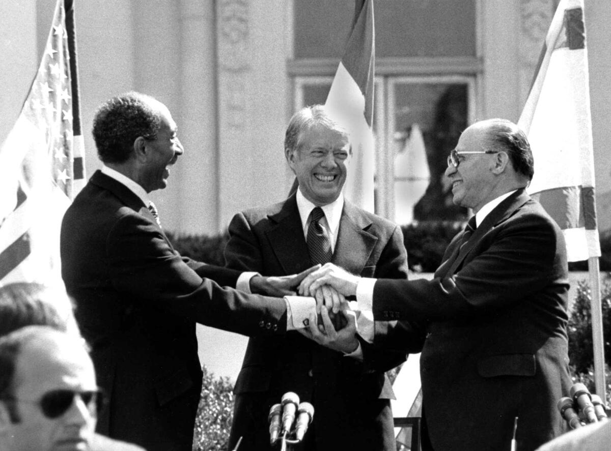 A black-and-white photo of Anwar Sadat, Jimmy Carter and Menachem Begin smiling, their hands piled together in the middle