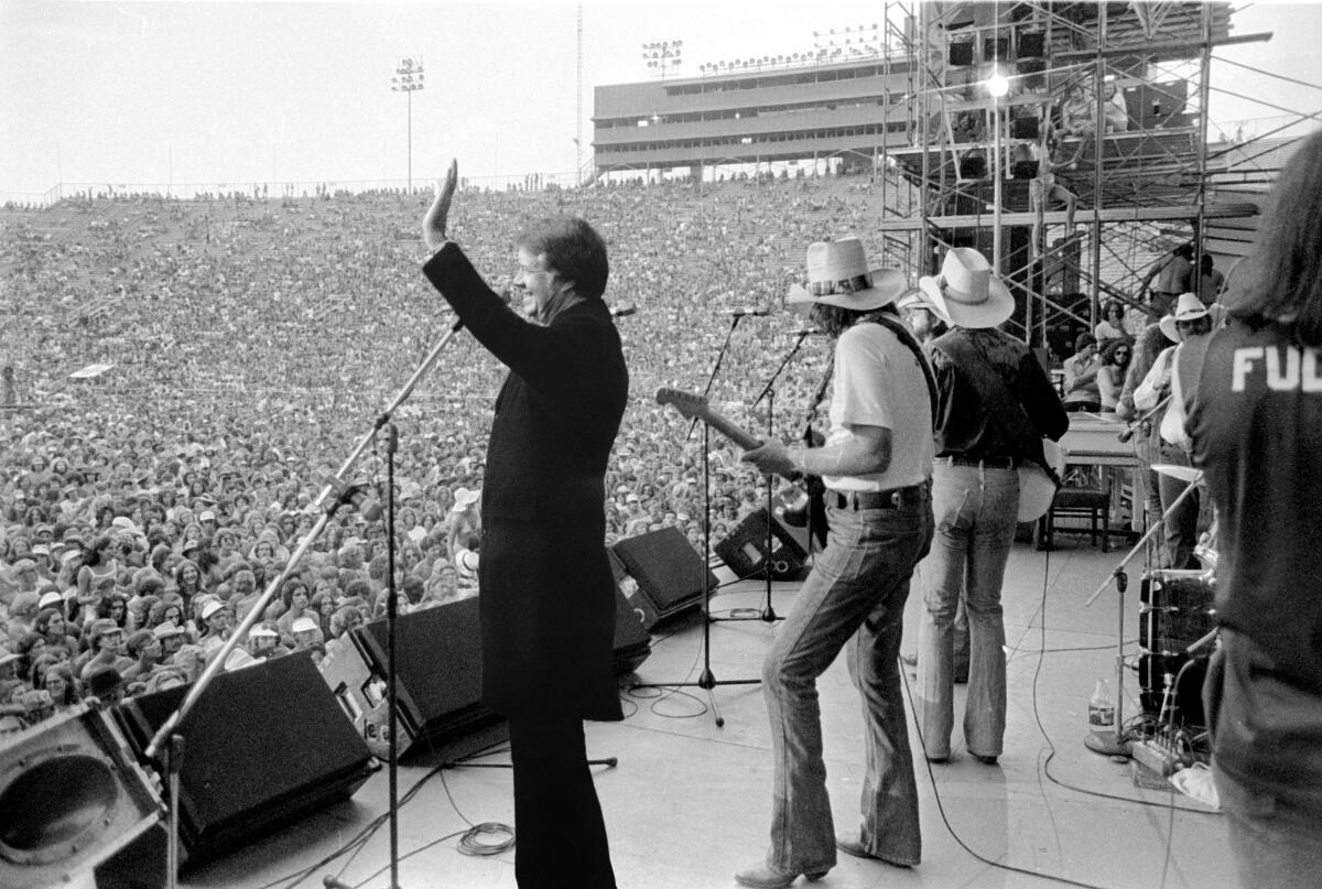 Jimmy Carter, onstage with Willie Nelson, waving to a large concert crowd.