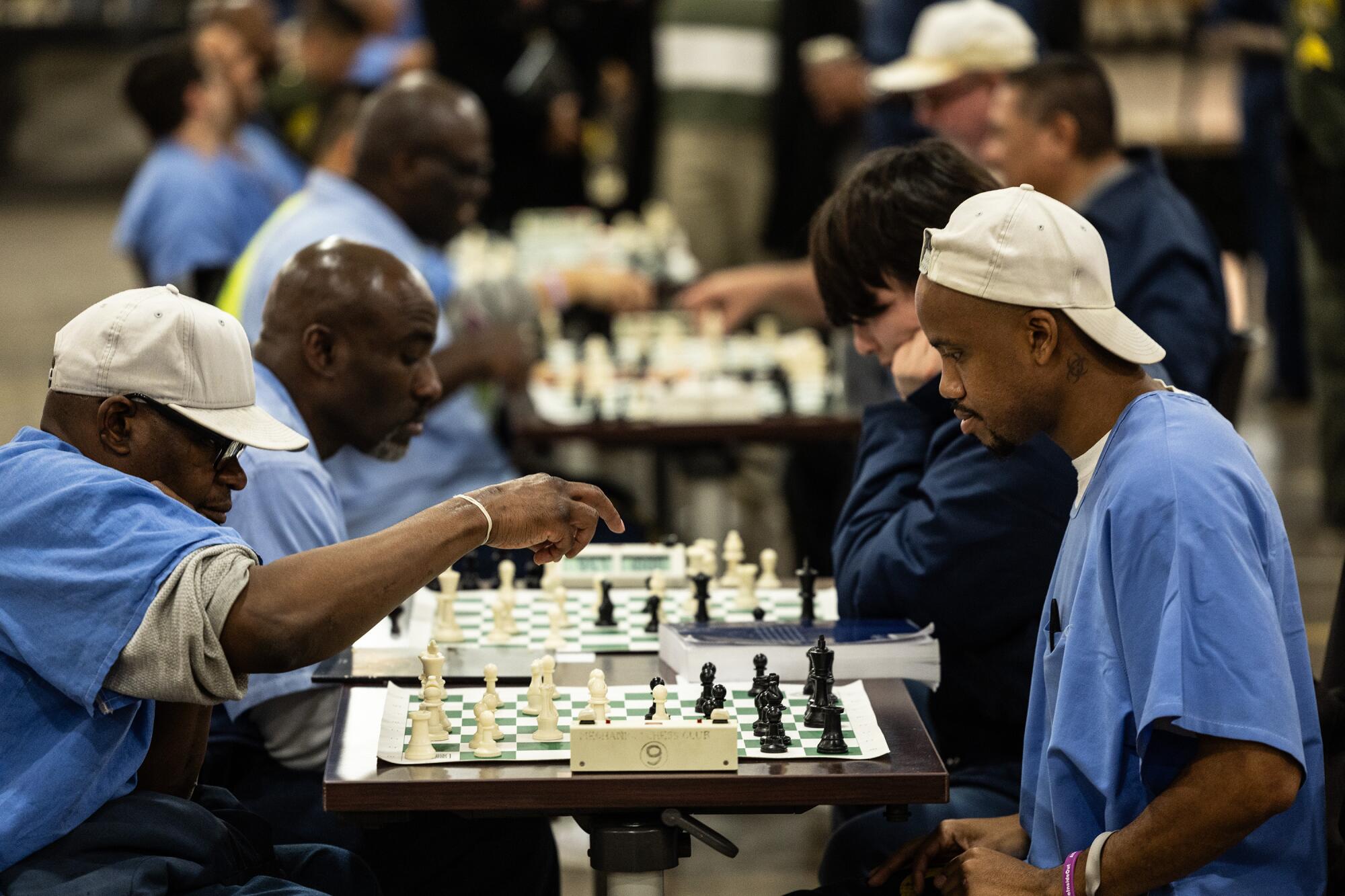 Detainees in blue prison garb play chess at a row of tables