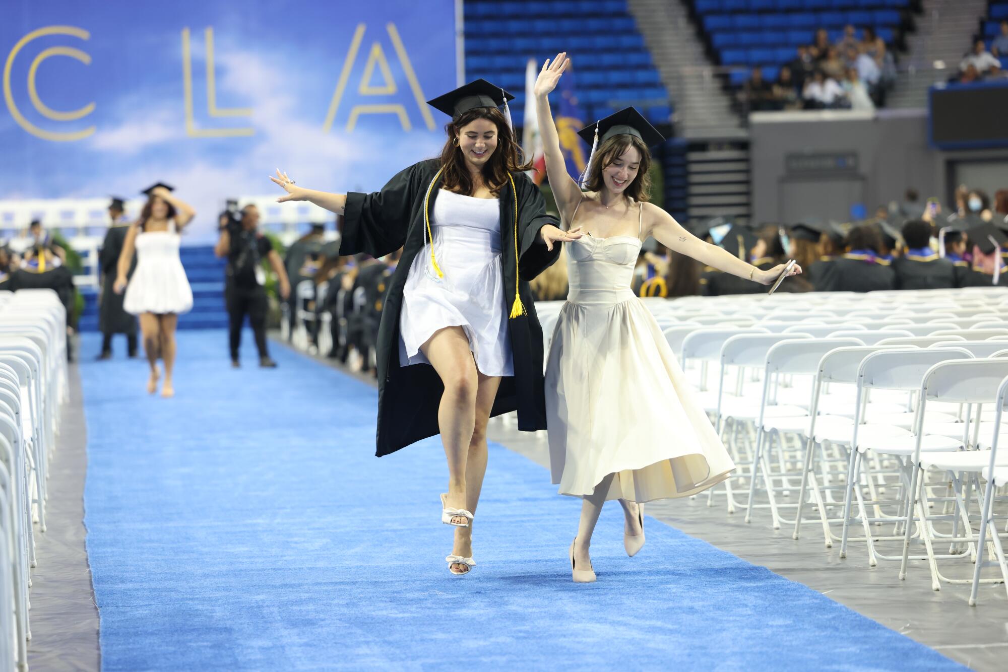 Two graduates in mortarboard hats dance down a blue carpet at UCLA commencement