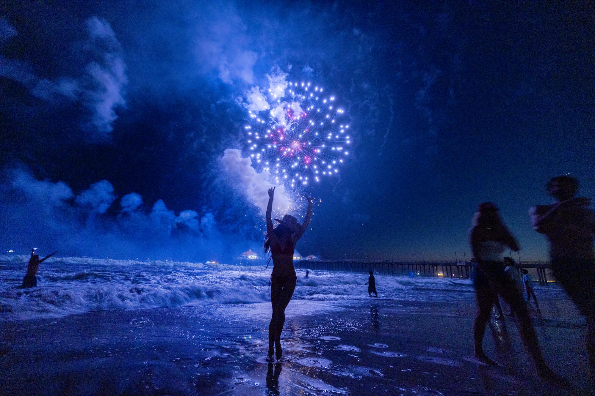 Beachgoers are in silhouette as an exploding firework casts blue light on the shore