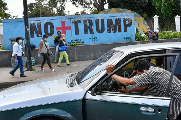 A person leans into an open car to push it down the street, in front of a sidewalk mural in Venezuela that reads "No + Trump"