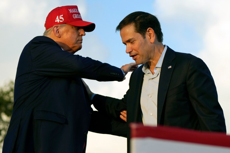 Donald Trump, wearing a red MAGA cap, reaches over to put a hand on Marco Rubio's shoulder