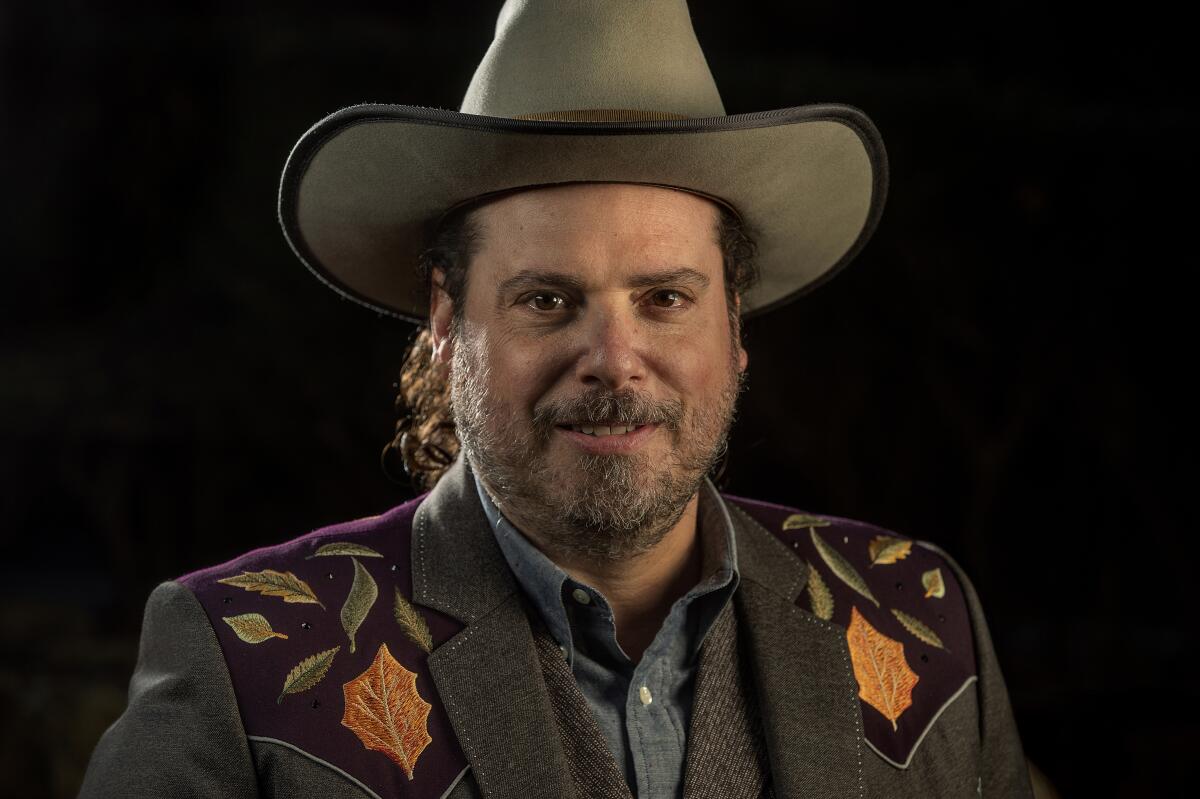 A bearded man wearing a cowboy hat and blazer with leaf designs at the shoulders