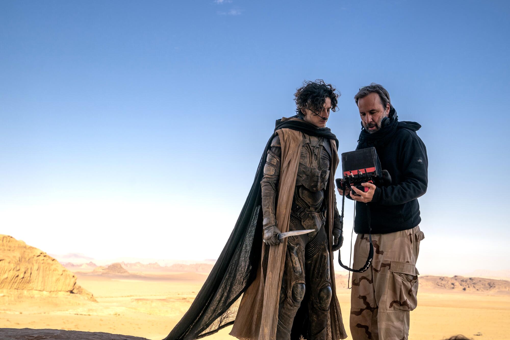 Two men, one in an elaborate costume, stand in the desert looking at an Ipad.