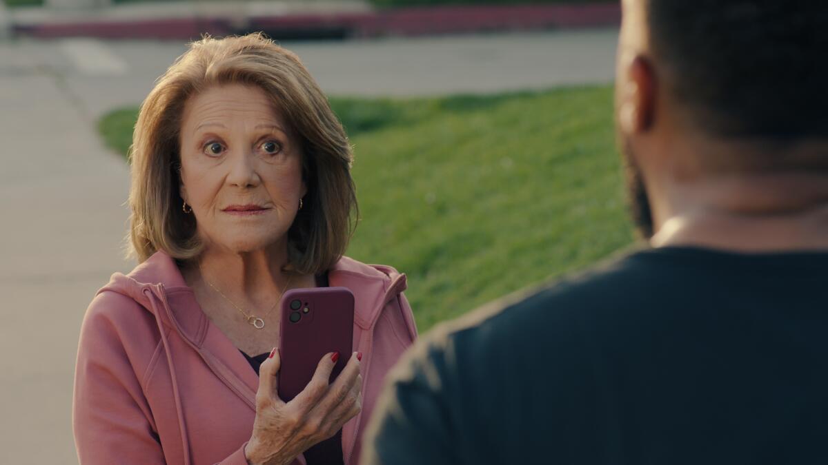 A woman in a pink sweatsuit holds up a phone while speaking with a man.