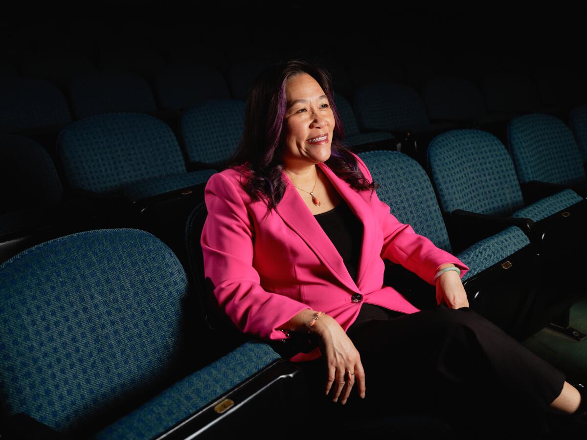 A woman in a pink blazer sits in a row of blue theater seats, smiling