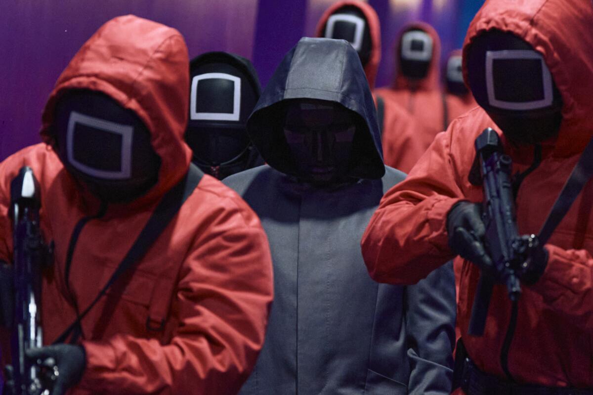 a man in a black hooded coat surrounded by men in pink hooded jumpsuits holding guns