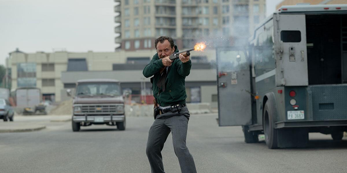 A man stands in the road and fires a shotgun.