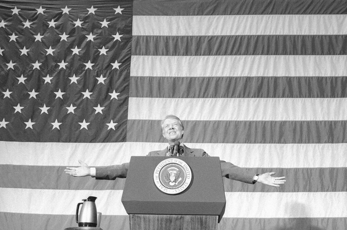 President Carter holds out his arms and smiles in front of a U.S. flag backdrop.