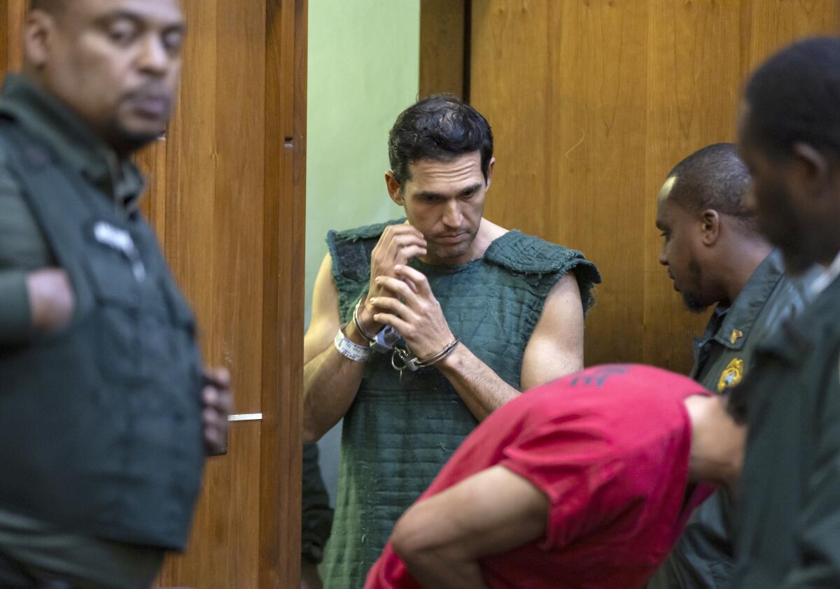 Two men in a courtroom surrounded by security personnel.