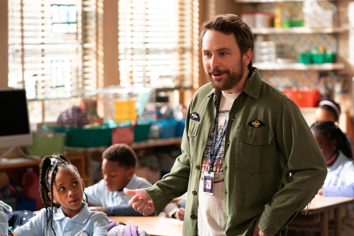 A man in a green jacket stands in a classroom as a child looks at him from her desk.