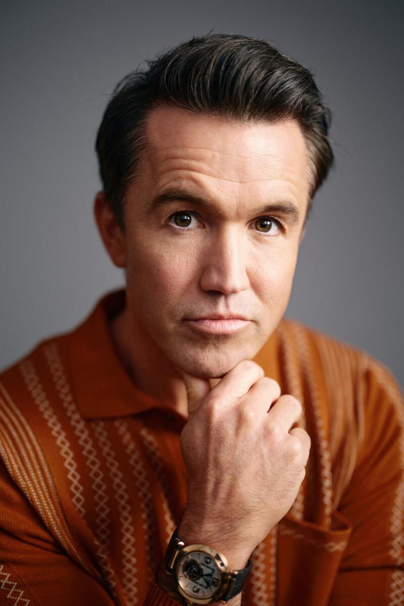 A close-up portrait of a man in an orange shirt holding his hand to his chin.