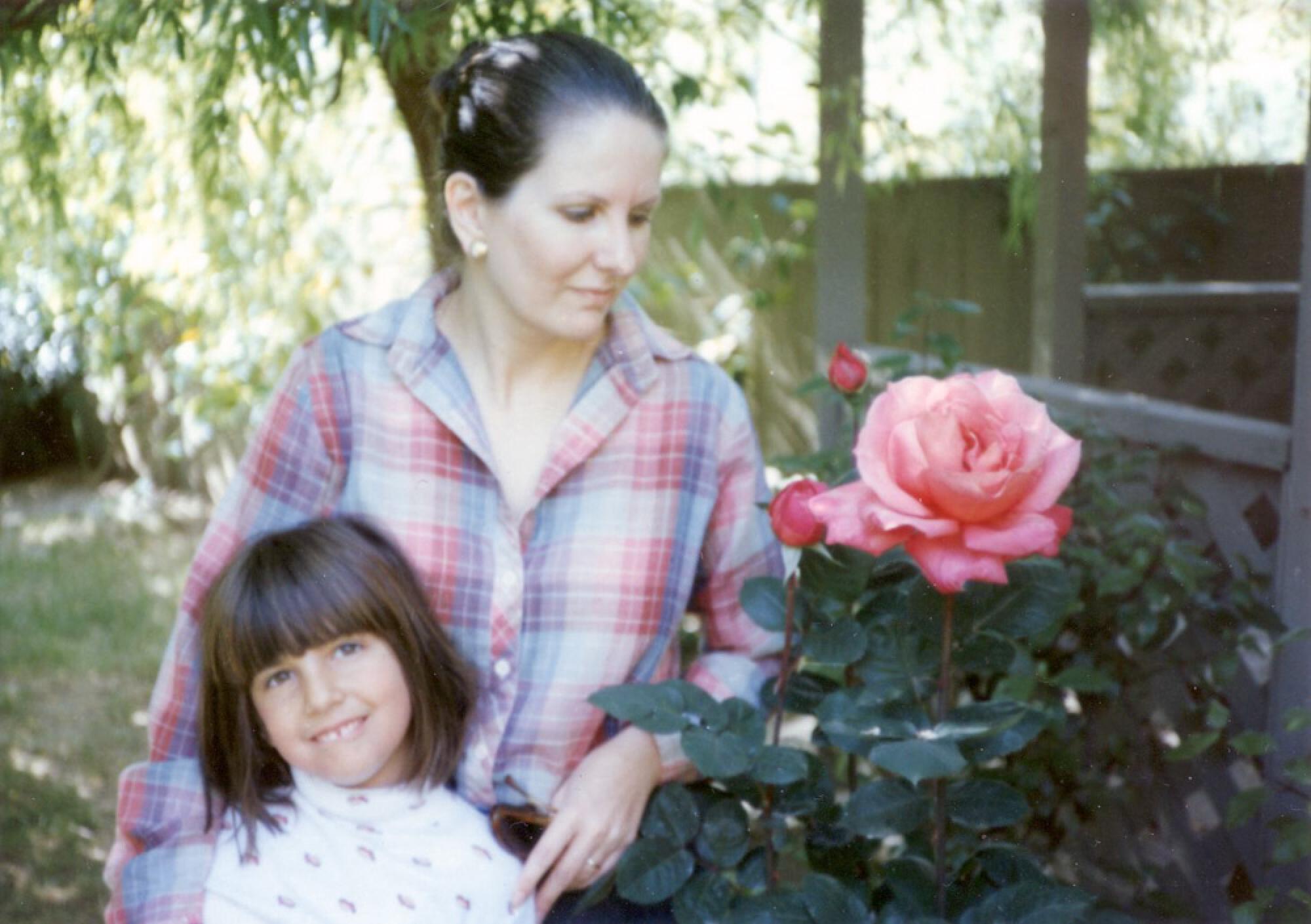 A woman with dark hair, in a plaid red-and-blue shirt, standing behind a young girl near a rosebush with pink blooms