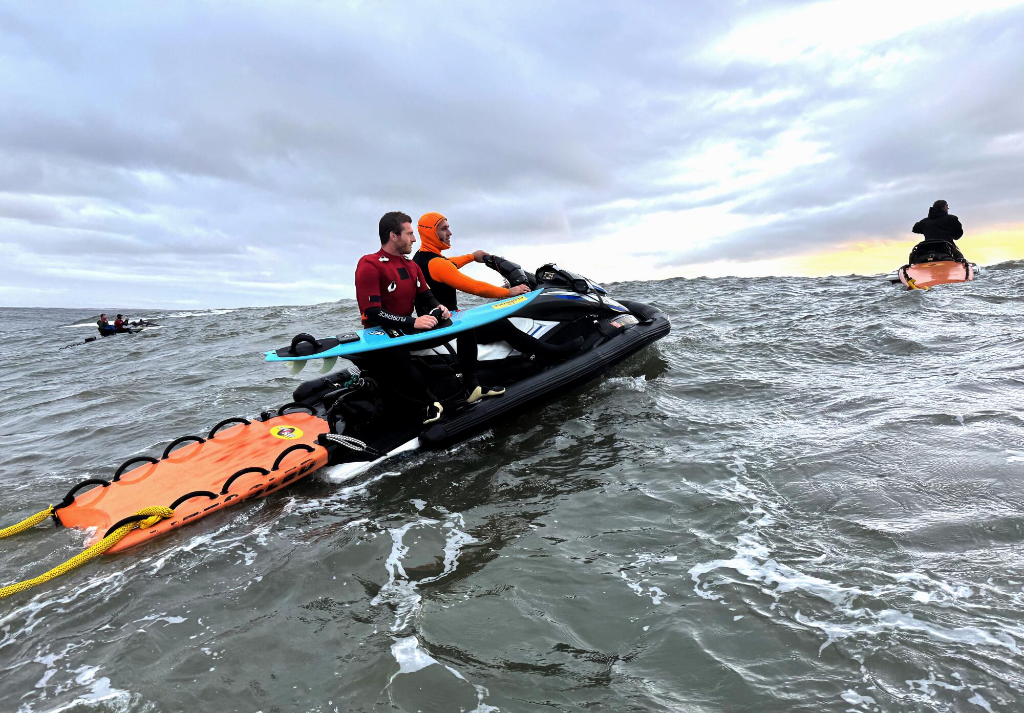 Alo Slebir, holding a turquoise surf board, hitches a ride on a jet ski at Mavericks Beach. 