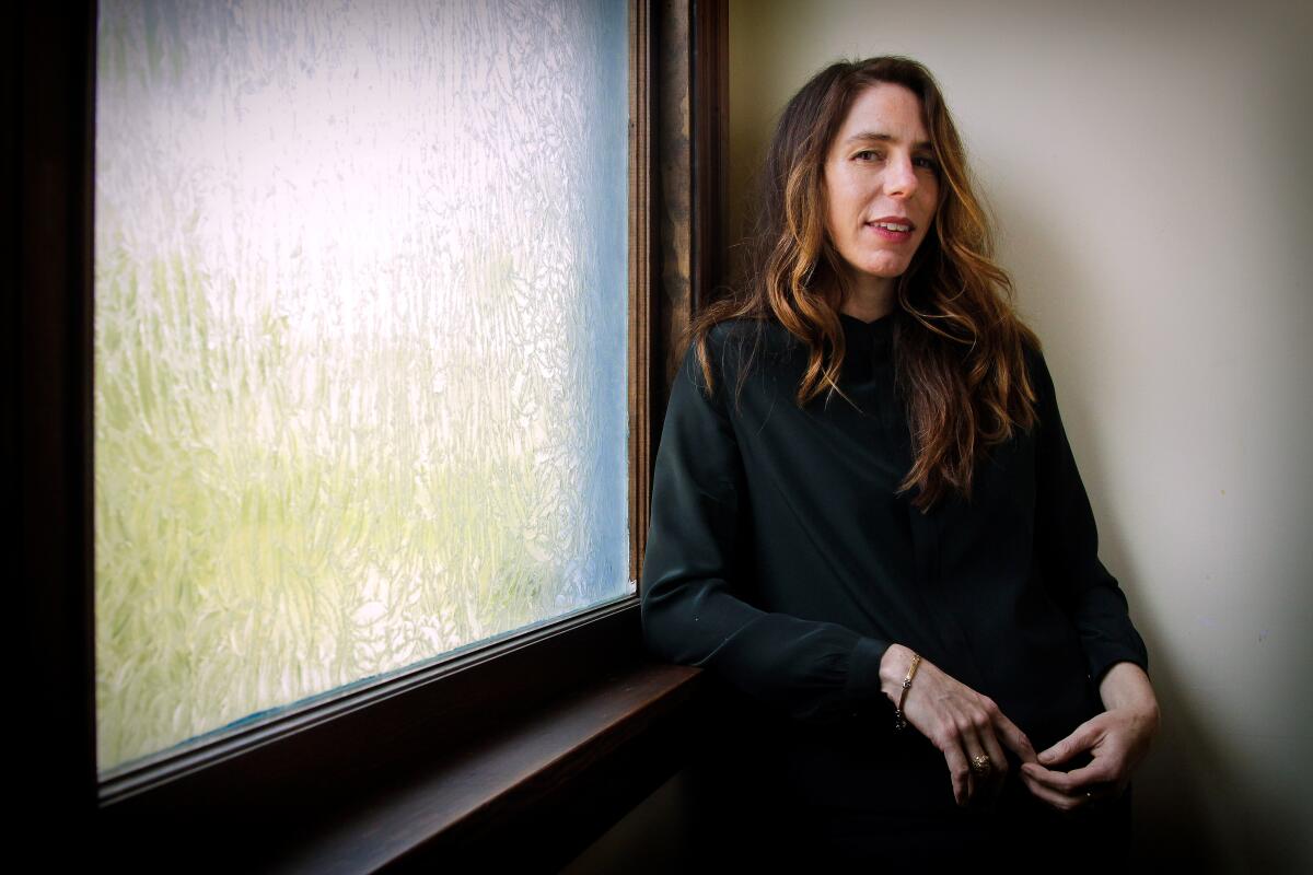 Novelist Rachel Kushner leans against a window sill next to a window.