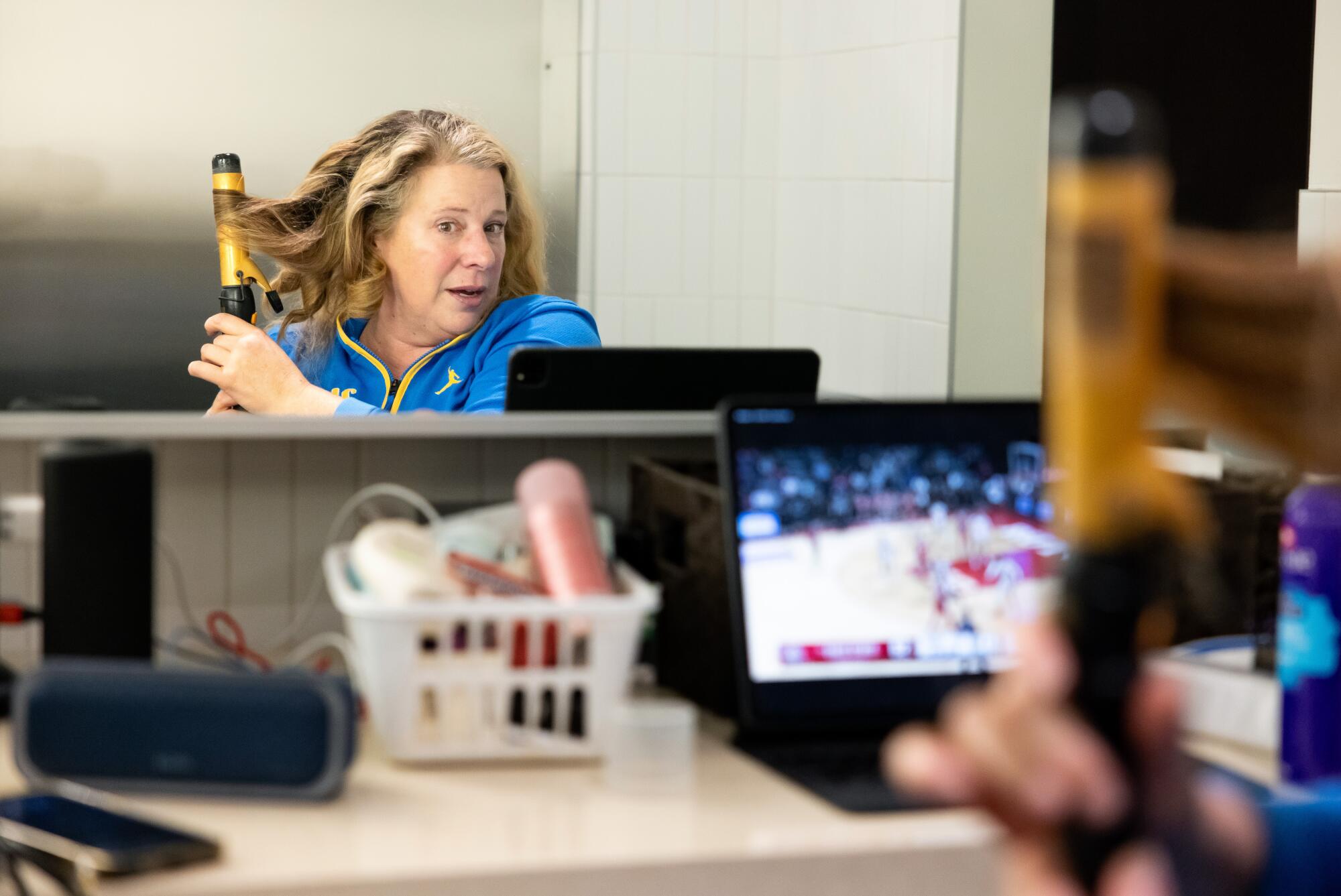 UCLA Women's Basketball Head Coach Cori Close curls her hair and watches a women's basketball game on her laptop.