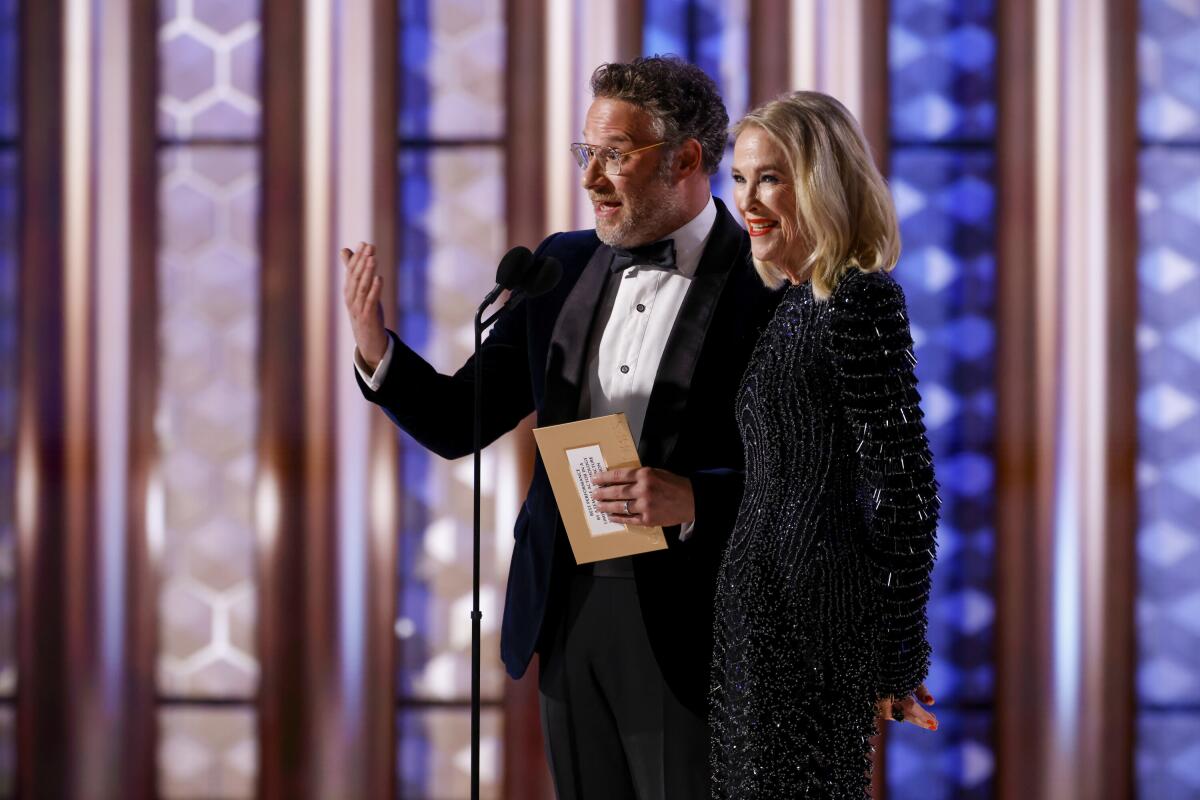 A man in a black tuxedo standing next to a woman in a black long sleeve gown on a stage.