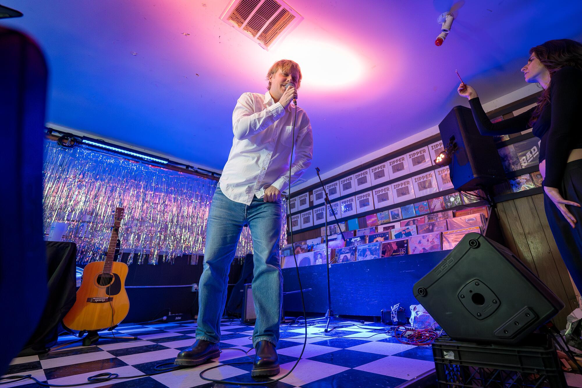 Comedian John Early sings on a stage, holding a microphone, in front of silver tinsel.