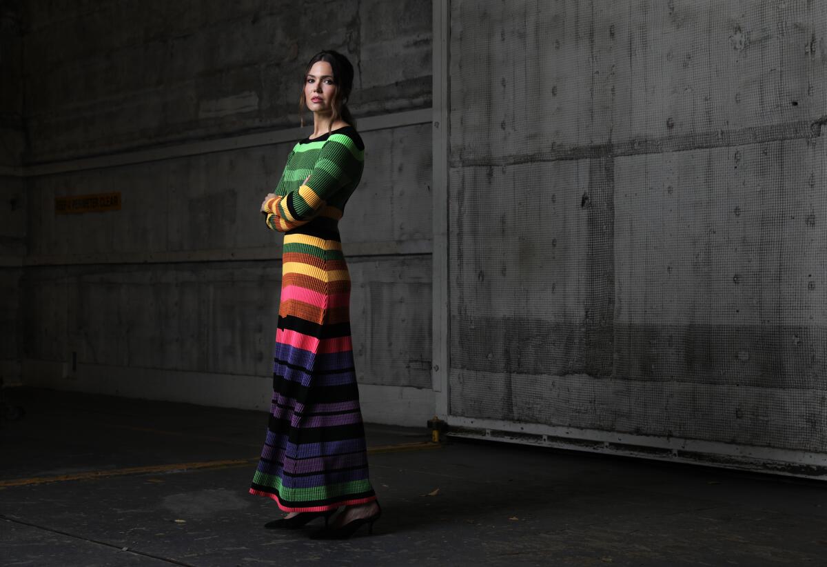 Mandy Moore stands with her arms crossed while posing for a portrait in a colorful, striped dress