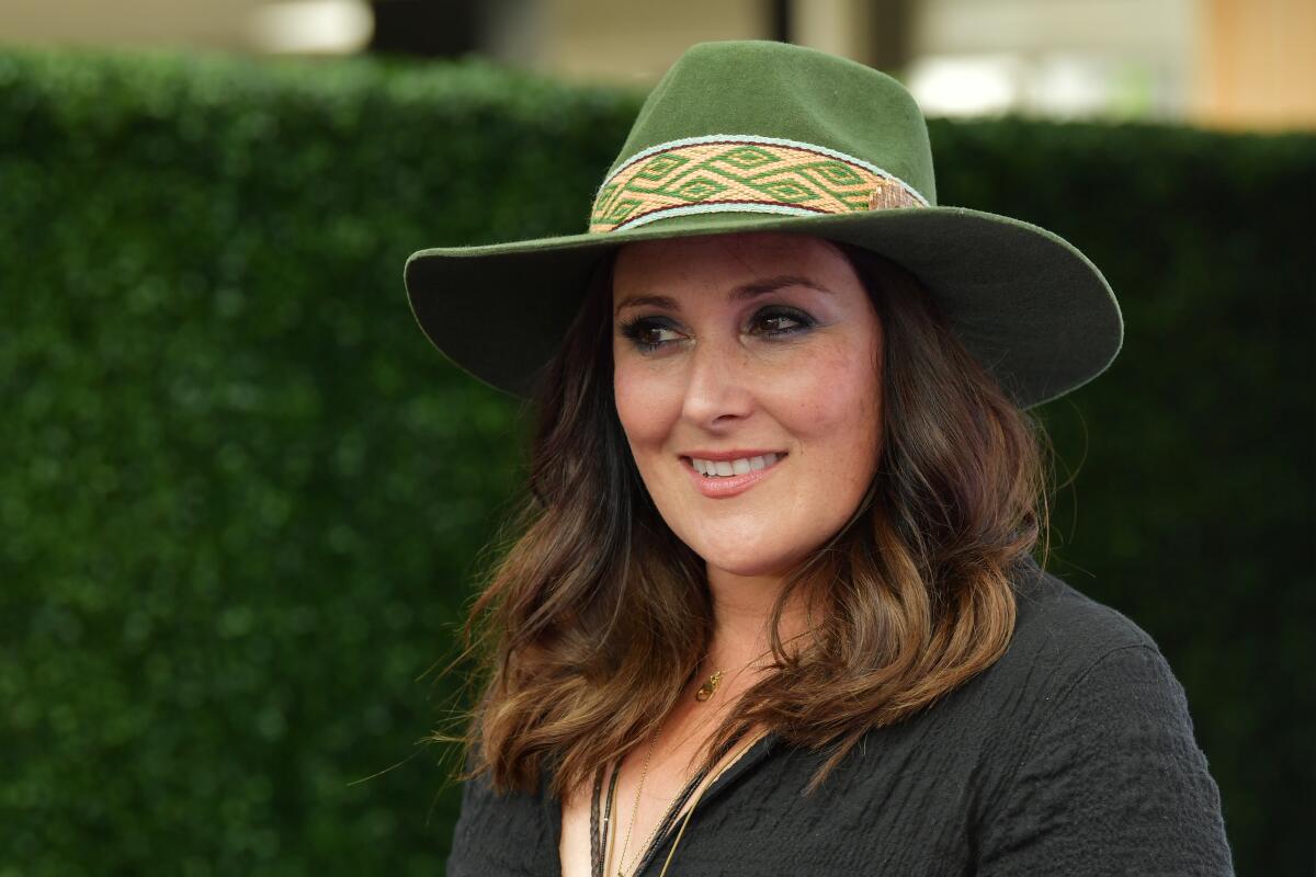 Ricki Lake poses in a green, wide-brimmed hat in front of a hedge