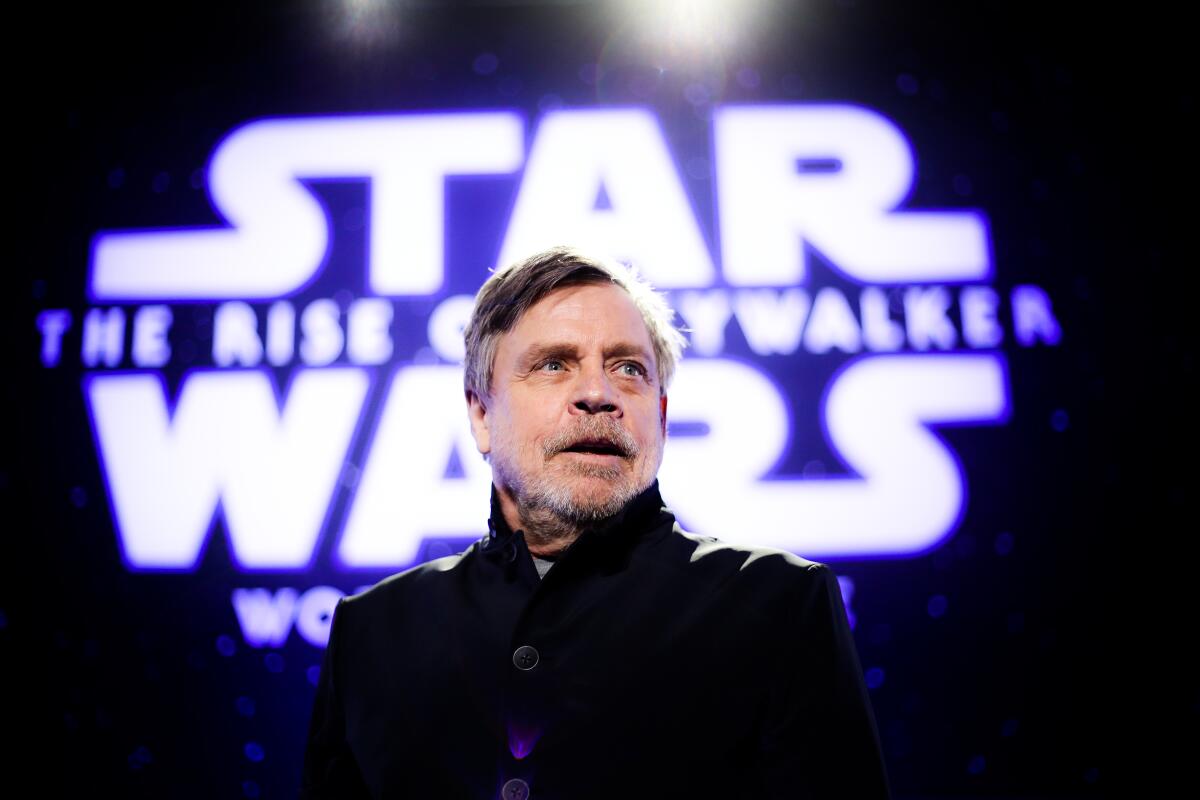 Mark Hamill speaking in front of a Star Wars logo in a black button-up jacket