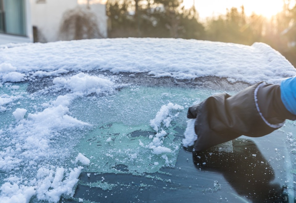 A gloved hand clears snow and ice from a car windshield.