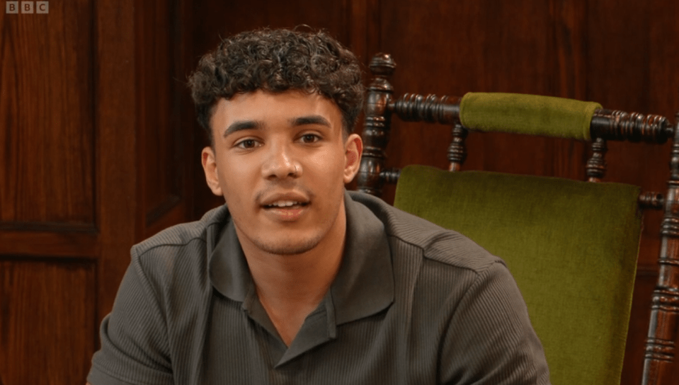 A young man with curly hair sits in a chair and speaks.