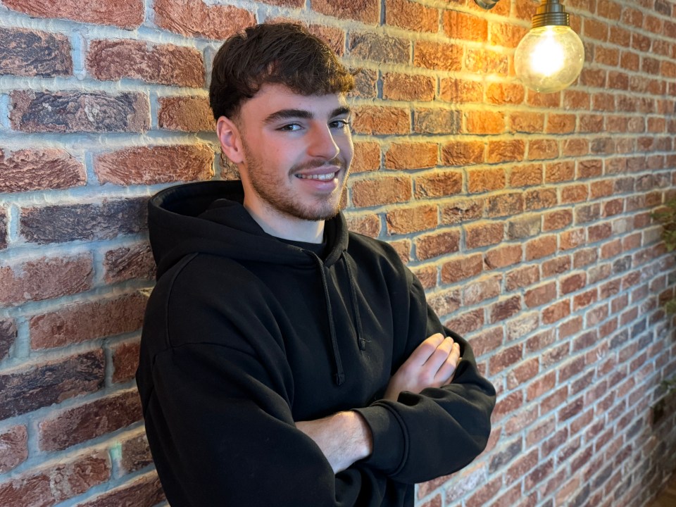 Young man in black hoodie with arms crossed, standing against brick wall.