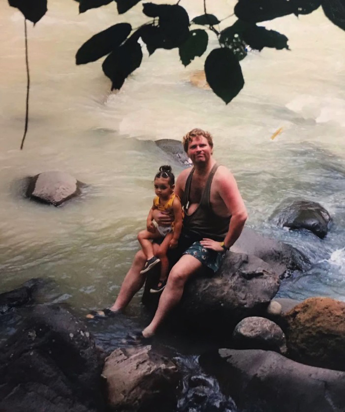 A man and a young child sitting on rocks in a shallow stream.