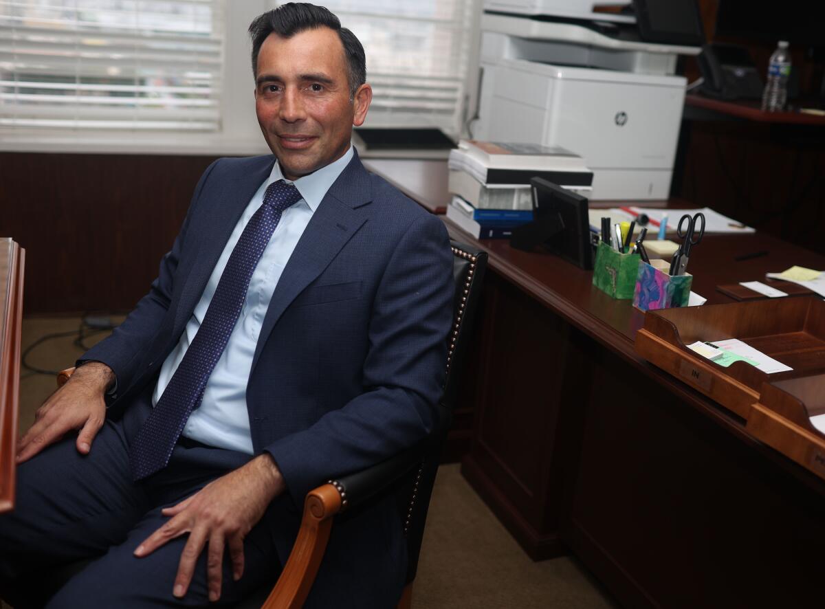 A smiling man with dark hair, in a dark suit and tie, seated in front of a desk with a white machine and office supplies