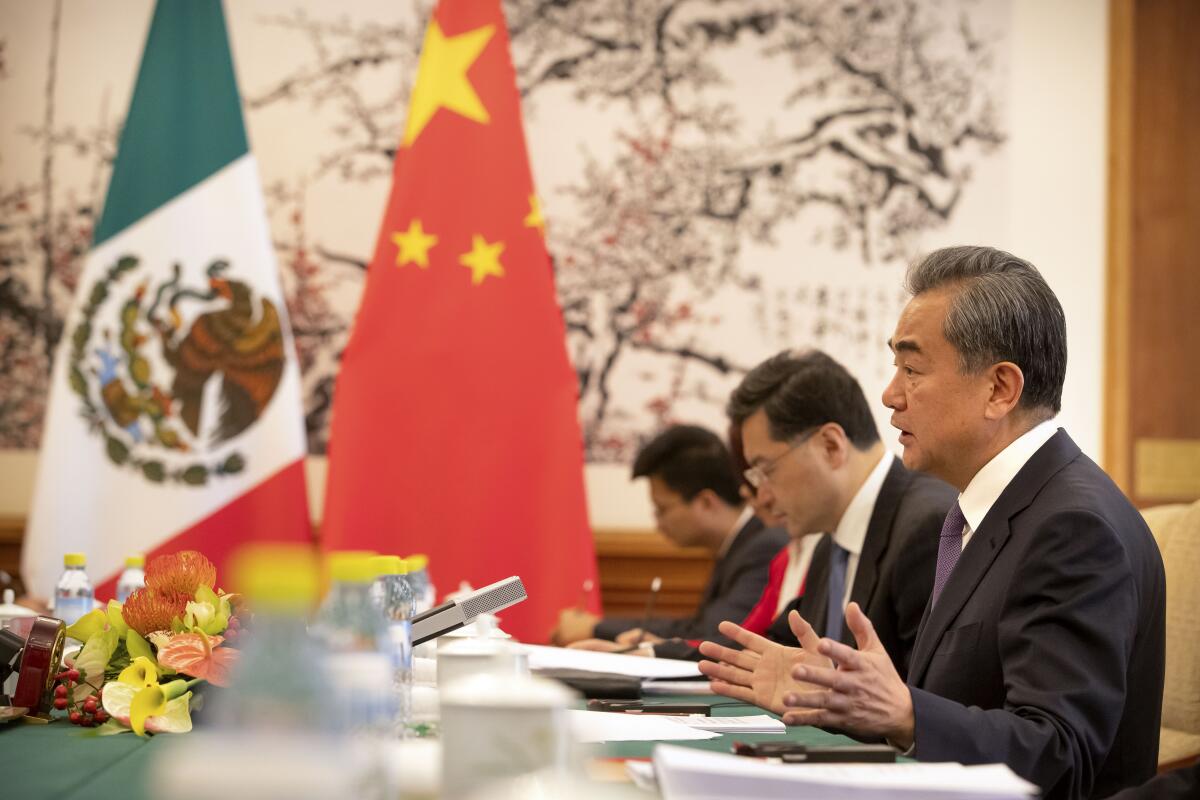 A man in a suit and tie gestures with his hands while seated at a table near a green, white and red flag and another in red