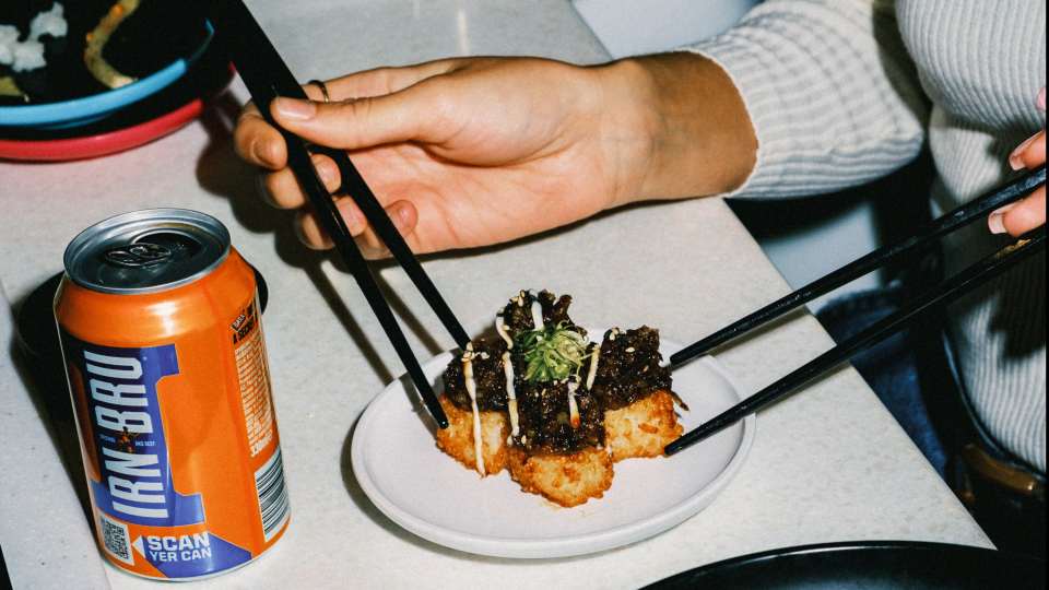 Person using chopsticks to eat a small dish of food next to a can of Irn-Bru.