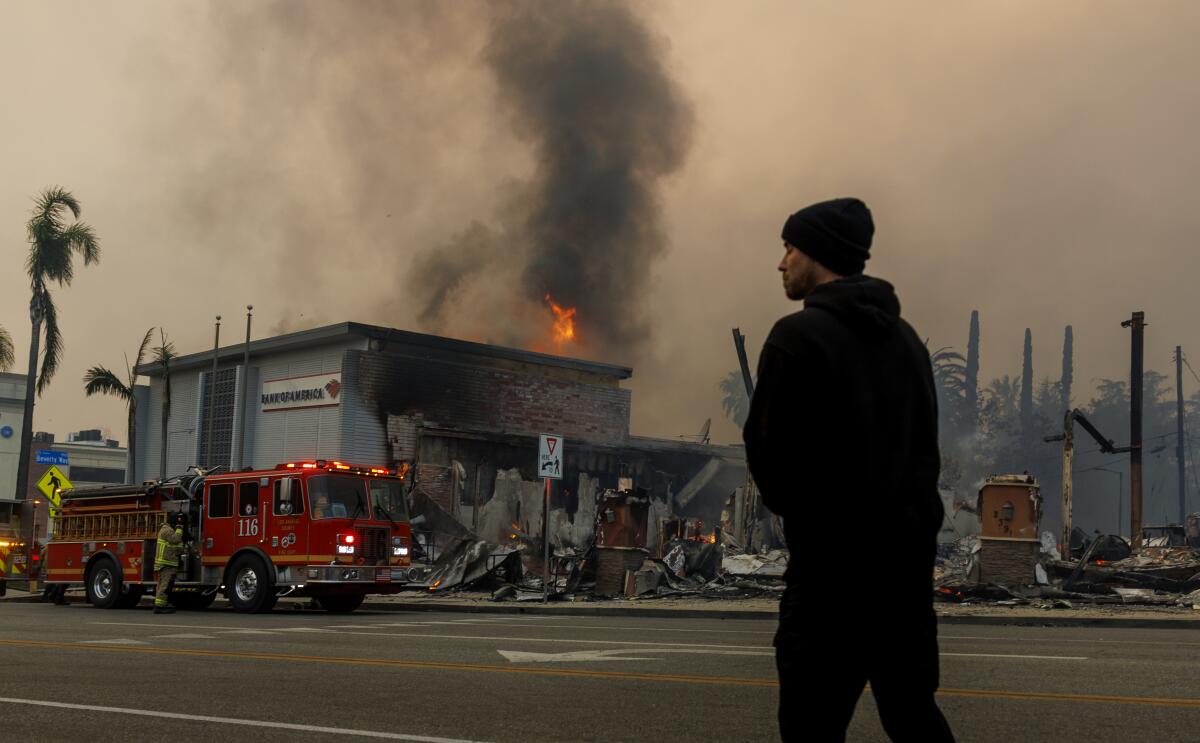 Flames rise from the roof of a building as a firetruck sits out front with red lights on.