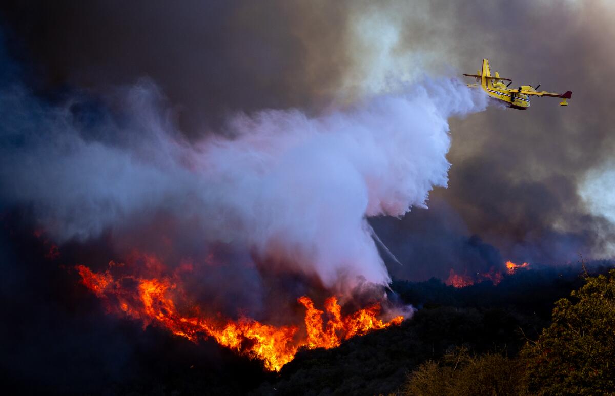 A yellow plane drops water on a fire in the hills below.