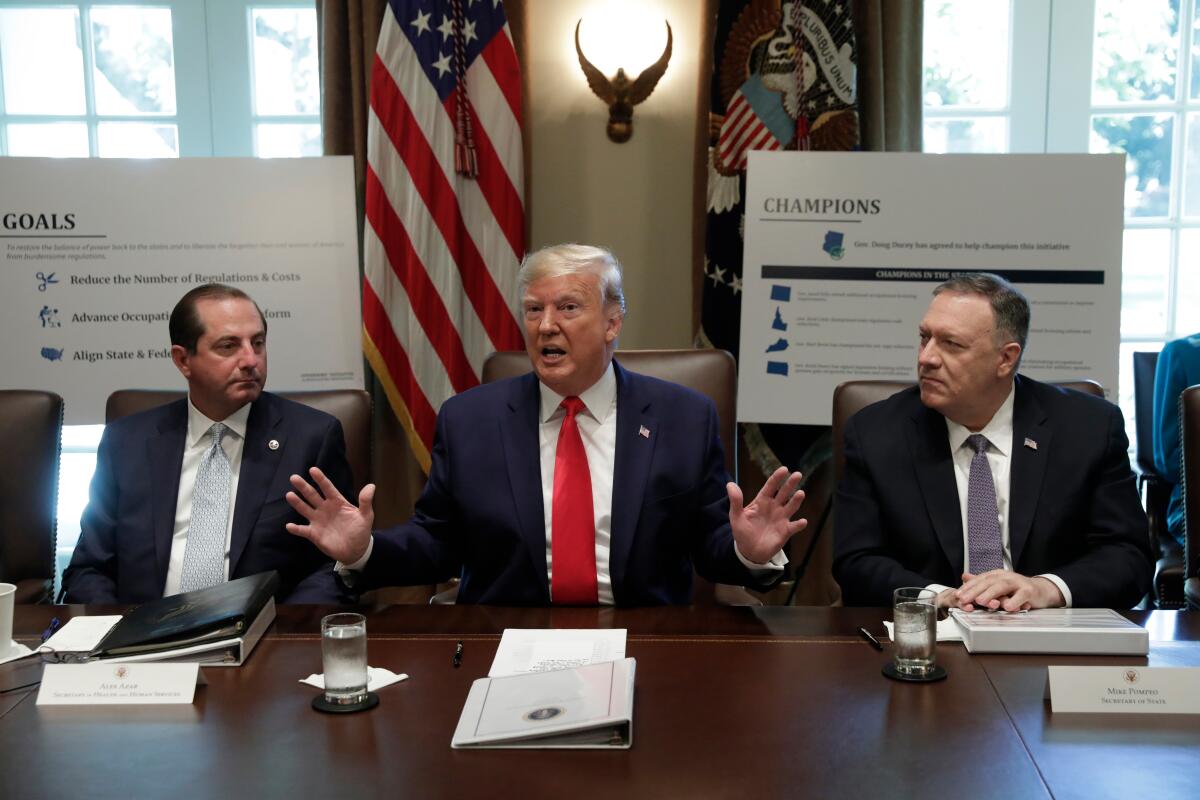 A man in dark suit and red tie gestures with his hands while speaking, seated at a desk with two men, also in suits and ties