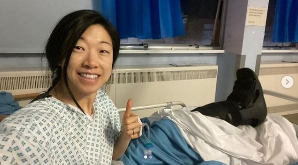 Woman in hospital gown giving thumbs up, leg in cast.