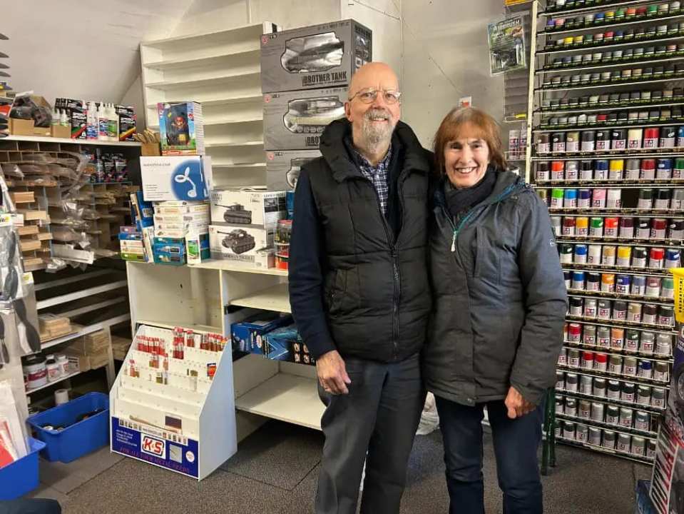 Two people stand in a model shop, surrounded by model kits and paints.