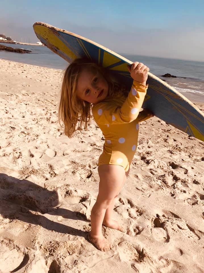 Toddler in yellow polka dot swimsuit carrying a boogie board on a sandy beach.