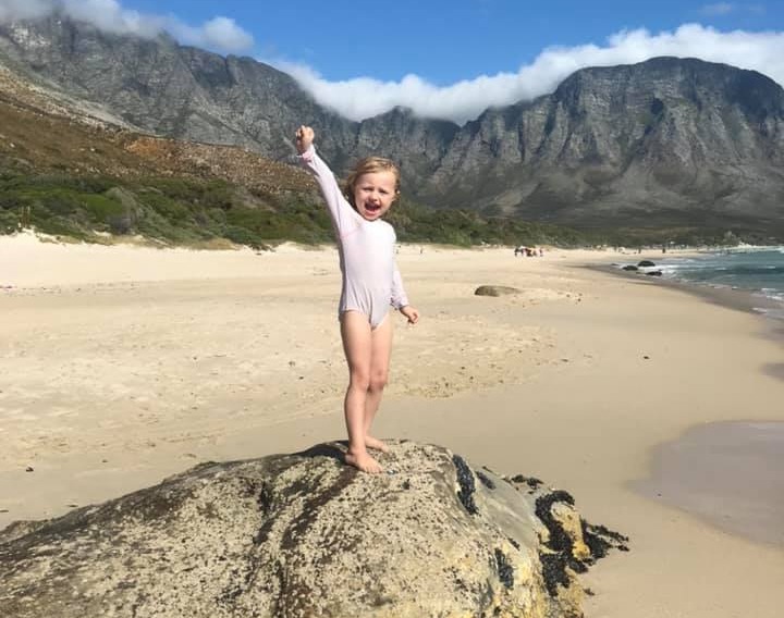 Girl standing on rock at beach with mountains in background.